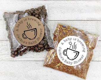 May include: Two clear plastic bags with white circular labels. The labels have black text that reads "A baby is brewing Jackie's Baby Shower". One bag contains coffee beans and the other contains loose leaf tea.