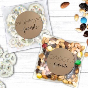 Two clear bags are on a white wooden surface. One contains yogurt-covered pretzels labeled "Mommy's favorite," and the other holds mixed nuts and candies labeled "Daddy's favorite." Nearby, loose pretzels and trail mix are scattered.