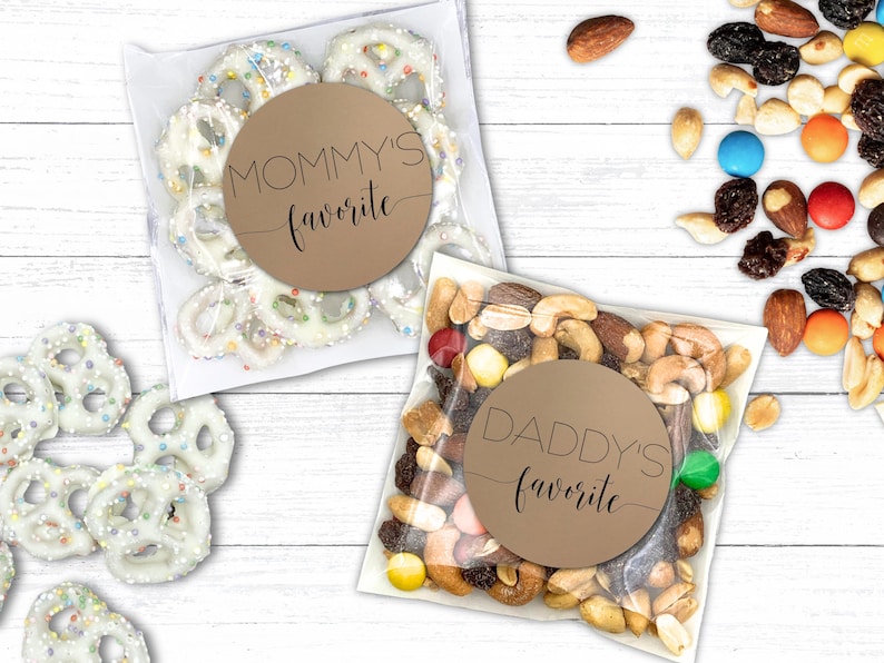Two clear bags are on a white wooden surface. One contains yogurt-covered pretzels labeled "Mommy's favorite," and the other holds mixed nuts and candies labeled "Daddy's favorite." Nearby, loose pretzels and trail mix are scattered.