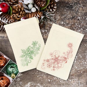 K&ouml;nnte beinhalten: Zwei wei&szlig;e Stoffgeschenkt&uuml;ten mit Kordelzug. Eine Tasche hat ein gr&uuml;nes Schneeflockenmuster und die andere ein rotes Schneeflockenmuster.