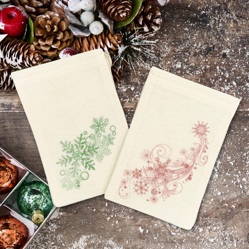 K&ouml;nnte beinhalten: Zwei wei&szlig;e Stoffgeschenkt&uuml;ten mit Kordelzug. Eine Tasche hat ein gr&uuml;nes Schneeflockenmuster und die andere ein rotes Schneeflockenmuster.