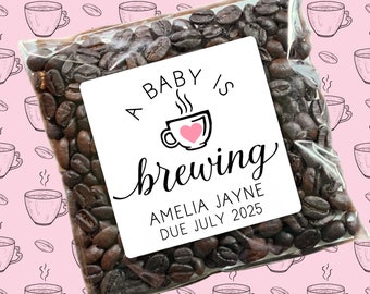 May include: A white favor bag filled with coffee beans and a white sticker that says "A baby is brewing Amelia Jayne Due July 2025". The sticker has a pink heart in a coffee cup.