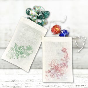 K&ouml;nnte beinhalten: Zwei kleine wei&szlig;e Stoffgeschenkt&uuml;ten mit Kordelzug. Eine Tasche hat ein gr&uuml;nes Schneeflockenmuster und die andere ein rosafarbenes Schneeflockenmuster. Die Taschen sind mit verschiedenen Schokoladenbonbons gef&uuml;llt.