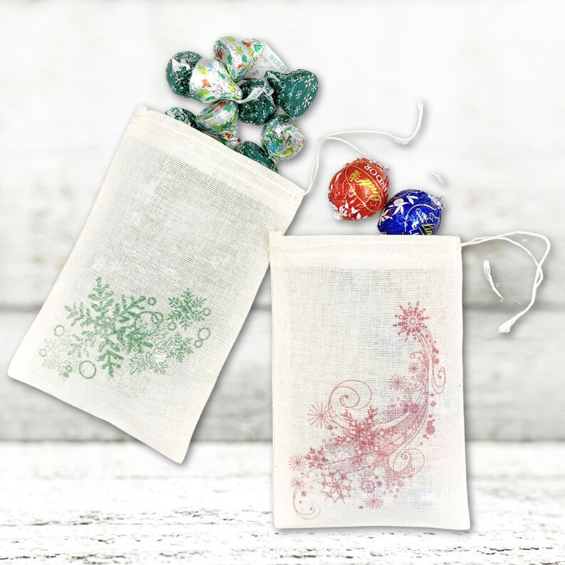 K&ouml;nnte beinhalten: Zwei kleine wei&szlig;e Stoffgeschenkt&uuml;ten mit Kordelzug. Eine Tasche hat ein gr&uuml;nes Schneeflockenmuster und die andere ein rosafarbenes Schneeflockenmuster. Die Taschen sind mit verschiedenen Schokoladenbonbons gef&uuml;llt.