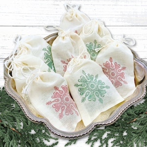 May include: A collection of small, off-white drawstring bags with snowflake designs in red and green, arranged on a decorative silver tray. The tray is surrounded by evergreen branches, creating a festive holiday theme.