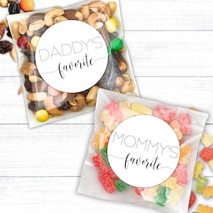 Two clear plastic bags filled with treats. One bag contains gummy bears with a round white sticker that says ‘MOMMY’S favorite'. The other bag contains trail mix with a round white sticker that says ‘DADDY’S favorite'.  Design printed in black.  MOMMY and DADDY in thin modern all caps.  “Favorite” in modern script spanning entire label.