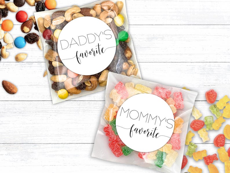 Two clear plastic bags filled with treats. One bag contains gummy bears with a round white sticker that says ‘MOMMY’S favorite'. The other bag contains trail mix with a round white sticker that says ‘DADDY’S favorite'.  Design printed in black.  MOMMY and DADDY in thin modern all caps.  “Favorite” in modern script spanning entire label.