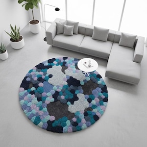 May include: A round, textured rug with a colorful, abstract design. The rug features a pattern of various shades of blue, gray, and purple. A gray sectional sofa and a small white table are also in the room. The rug is a decorative element for a living room.