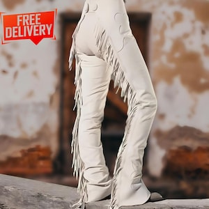 May include: Cream-colored leather chaps with long fringe detailing along the sides and bottom. The chaps feature decorative cutouts and are shown from the waist down. The words "FREE DELIVERY" are in a red box in the upper left corner.