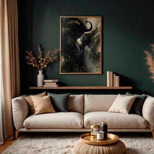 May include: A framed artwork depicting a dark, abstract figure embracing a lighter figure. The artwork hangs above a beige sofa with various throw pillows. A gold tray with candles and a small ottoman are in front of the sofa.
