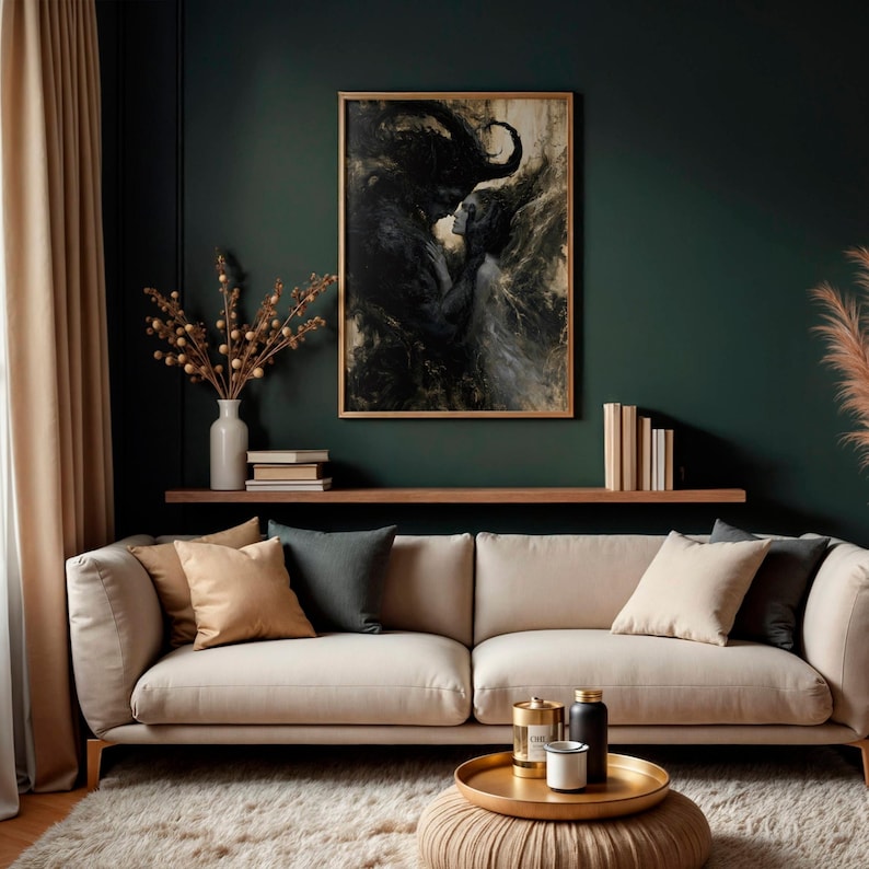 May include: A framed artwork depicting a dark, abstract figure embracing a lighter figure. The artwork hangs above a beige sofa with various throw pillows. A gold tray with candles and a small ottoman are in front of the sofa.