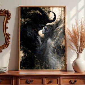 May include: A framed art print depicting a dark, romantic scene. A dark, shadowy figure embraces a woman with flowing hair. The artwork is in shades of black, gray, and gold, with a wooden frame. The print is displayed on a wooden dresser.