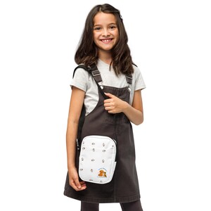 Könnte beinhalten: Eine weiße Umhängetasche mit einem sich wiederholenden Muster aus kleinen braunen Blumen und einer Grafik von zwei braunen Teddybären. Die Tasche wird von einer Person getragen, die ein braunes Latzkleid und ein graues T-Shirt trägt.