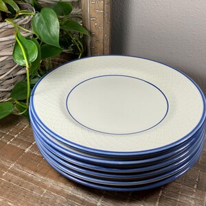 May include: A stack of six white ceramic plates with a blue border and a textured, crosshatch pattern. The plates have a cream-coloured centre and are stacked on a wooden surface. A plant and mirror are in the background.