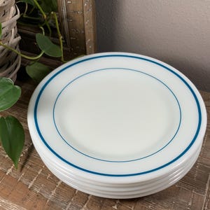 May include: A stack of white ceramic plates with a blue border. The plates are stacked on a wooden surface. The plates are a classic design, suitable for everyday use.