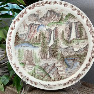 May include: A round ceramic decorative plate featuring a detailed illustration of Yosemite National Park. The design includes labeled landmarks: Half Dome, Bridal Veil Fall, El Capitan, Yosemite Falls, Tunnel Tree, Three Brothers, and Mirror Lake.