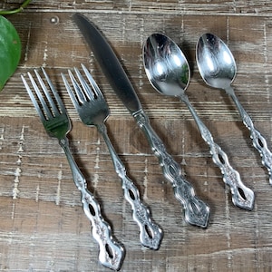 May include: A set of four stainless steel flatware pieces including two forks, one dinner knife, and two spoons. Each piece features a polished silver finish and a decorative handle with an intricate embossed floral pattern and a cutout design.