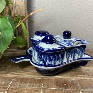 May include: A blue and white ceramic condiment server with three lidded compartments and a serving tray. The server features a grape vine design and is placed on a wooden surface.