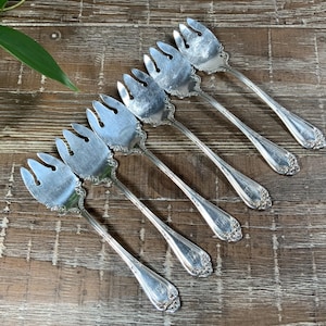 May include: Six silver-colored serving forks with three tines each are arranged diagonally on a wooden surface. The forks have ornate handles and are likely vintage or antique. The background is a rustic wooden table.