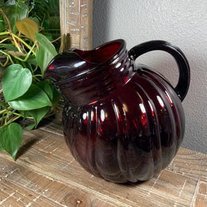 May include: A ruby red glass pitcher with a ribbed design. The pitcher has a rounded body, a curved handle, and a spout for pouring. The pitcher is sitting on a wooden surface.