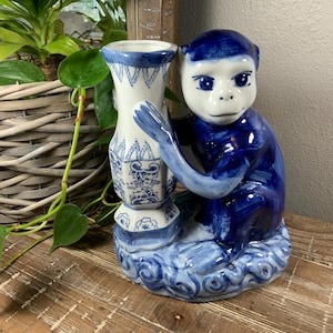 May include: A blue and white ceramic monkey figurine holding a matching vase. The monkey is sitting on a base with a wave design. The vase has a white background with blue patterns. The figurine is on a wooden surface.
