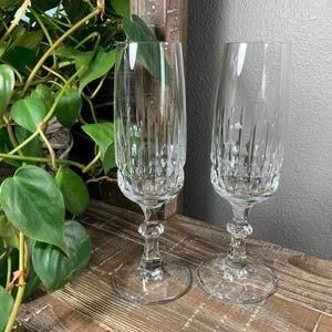 May include: Two clear, elegant champagne flutes with intricate vertical and circular cut glass patterns. The flutes have a classic design with a long stem and a wide base, perfect for special occasions. The glasses are displayed on a wooden surface.