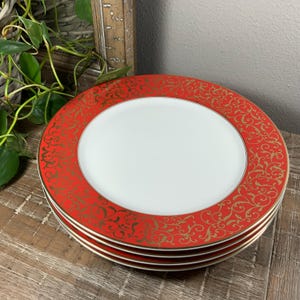May include: A stack of four round dinner plates featuring a solid white center and a wide red border. The red rim is decorated with an intricate, repeating gold filigree scroll pattern. Each plate has a thin gold line along the inner edge.