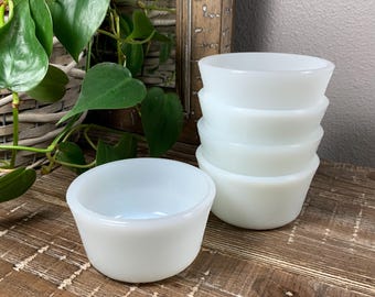 May include: A stack of five white, round, small bowls with a slightly flared rim. One bowl is separate, in front of the stack. The bowls are made of a smooth, opaque material. The bowls are on a wooden surface.