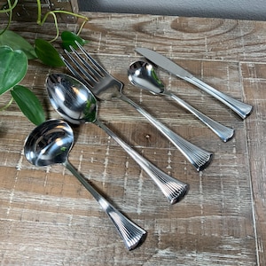 May include: A set of polished silverware including a fork, spoon, knife, and two serving spoons. The silverware has a fluted design on the handles and is arranged on a wooden surface. The set is ideal for formal dining.
