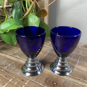 May include: Two cobalt blue glass goblets with a silver, ridged base. The glasses have a wide, open bowl shape and sit on a decorative, metallic base. The goblets are displayed on a wooden surface, with greenery in the background.