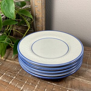 May include: A stack of six white ceramic plates with a blue rim and a textured, crisscross pattern. Each plate has a smaller, light gray center. The plates are stacked on a wooden surface.