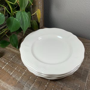 May include: A stack of four white ceramic plates with a scalloped edge design. The plates are stacked on a rustic wooden surface, with green leafy plants in the background. The plates have a simple, elegant design.