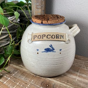 May include: A ceramic popcorn jar with a cork lid. The jar is off-white with a blue rim and handle. The word "POPCORN" is displayed on a banner. A blue rabbit and heart design is painted on the jar.