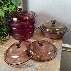May include: A collection of vintage glass cookware with lids. The set includes several round, ribbed dishes in shades of purple and amber. The lids have a central knob and a sunburst design. The cookware is arranged on a wooden surface.