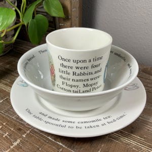 May include: A white ceramic children's tea set featuring a cup, bowl, and saucer. The cup and bowl have text from a children's story. The saucer has a tea cup illustration. The set is on a wooden surface.