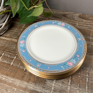 May include: A stack of white ceramic plates with a light blue floral border and gold trim. The floral design includes pink and blue flowers. The plates are stacked on a wooden surface.