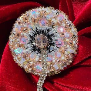 May include: A round, beaded brooch with a dandelion design. The brooch features iridescent sequins, clear and gold beads, and a central crystal. A dangling chain of crystals hangs from the bottom. The brooch is set against a red velvet background.