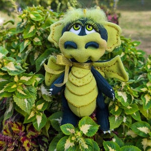May include: A plush dragon toy featuring a light green and dark green body with gold glitter accents on the wings and chest. The creature has large green eyes with long eyelashes, a tuft of light green hair, and a gold ribbon tied around its neck.