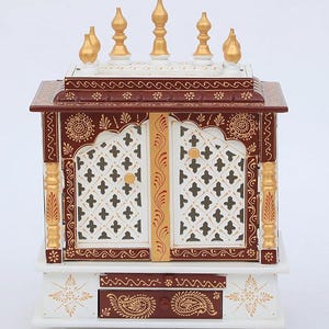 May include: A decorative wooden cabinet with intricate detailing. The cabinet features a white base, a dark brown top, and white doors with a lattice design. Gold accents and ornate patterns adorn the exterior, including the drawer and pillars. The cabinet is topped with gold finials.