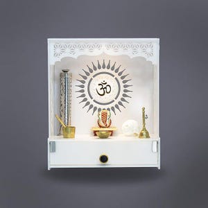 Puede incluir: Templo de madera blanca con un diseño decorativo de sol y el símbolo Om. El templo tiene un cajón con un pomo negro y contiene varios objetos religiosos, incluyendo una estatua, una campana y un recipiente.