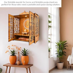 Puede incluir: Un mandir de madera plegable con intrincados dise&ntilde;os calados, montado en una pared blanca. El mandir tiene un estante y una peque&ntilde;a luz en el interior. La imagen tambi&eacute;n muestra dos macetas naranjas con plantas sobre una mesa de madera. El texto dice: Nuestro mandir plegable para el hogar y el soporte pooja plegable son perfectos.