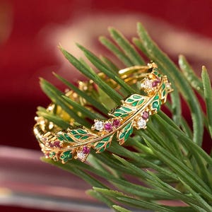 Peut inclure: Une bague dorée avec un motif de feuilles, ornée d'émail vert, de pierres précieuses rouges et blanches. La bague est présentée sur une branche de pin contre un fond rouge, suggérant un thème festif.