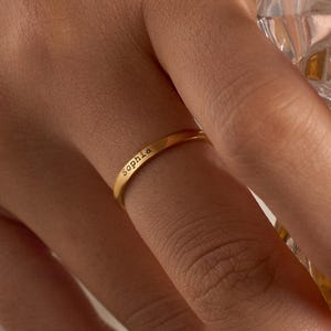 Peut inclure: Une bague dorée gravée du nom "Sophia". La bague est fine et portée au doigt. L'image est un gros plan, avec un verre de liquide ambré en arrière-plan.