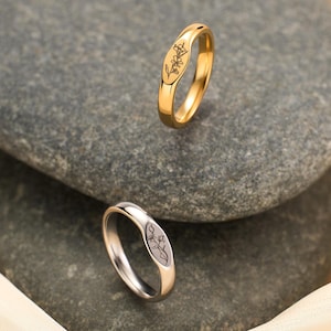 May include: Two rings, one gold and one silver, each featuring a floral design. The rings are displayed on a grey stone, with a soft, cream-coloured fabric in the background. The gold ring has a polished finish, the silver ring a matte finish.