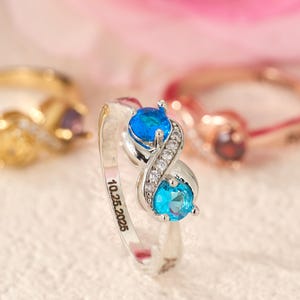 May include: A silver ring featuring two blue gemstones and small white stones, with the date "10.25.2025" engraved on the band. Two other rings are visible in the background: a gold ring with a yellow stone and a rose gold ring with a red stone.