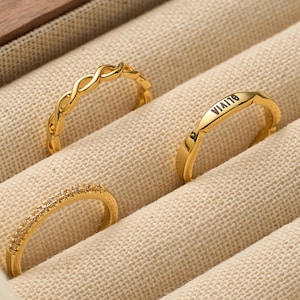 May include: Three gold rings presented in a jewellery box. One ring has an interwoven design, another is engraved with the name "Olivia", and the third is decorated with small, sparkling stones. The rings are set against a beige fabric.