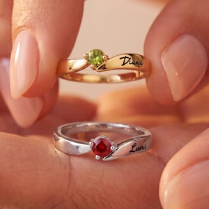 Peut inclure: Deux bagues, une en or et une en argent, sont tenues dans une main. La bague en or a une pierre verte et le nom "Diana" gravé. La bague en argent présente une pierre rouge et le nom "Luna".