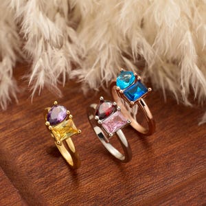 May include: Three rings with colourful gemstones. One ring features purple and yellow stones, another has red and pink stones, and the third has blue stones. The rings are displayed on a wooden surface.