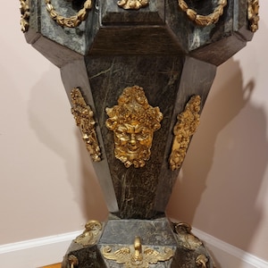 May include: A dark green and gold decorative pedestal with ornate gold accents. The pedestal features gold face and swan designs, and is made of a dark green stone-like material. The pedestal has a geometric shape and is likely used for display.