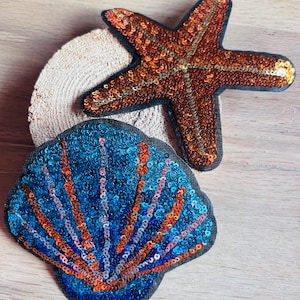 Könnte beinhalten: Gestickte Aufnäher mit einem Seestern und einer Muschel. Der Seestern ist braun mit goldenen Pailletten, die Muschel blau mit orangefarbenen und goldenen Pailletten. Ideal zum Aufnähen auf Kleidung oder Accessoires.
